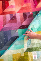 Colorful quilt with geometric patterns and a hand touching it, featuring the brand 'Quilty Love'.