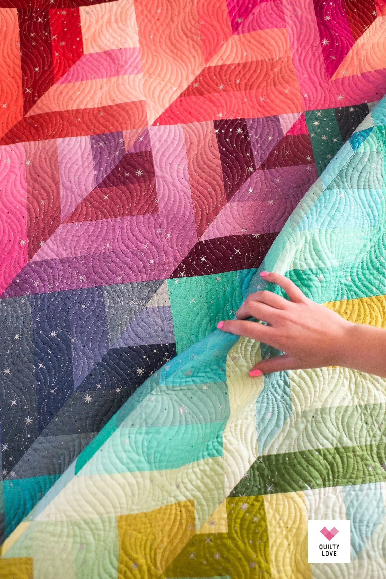 Colorful quilt with geometric patterns and a hand touching it, featuring the brand 'Quilty Love'.