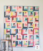 Colorful geometric quilt hanging on a wall with a small plant and stool in the foreground.
