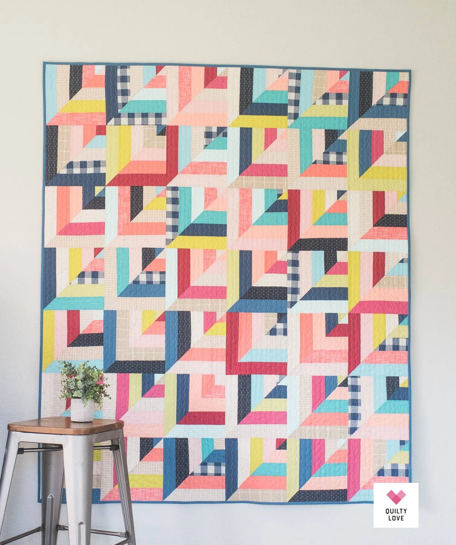 Colorful geometric quilt hanging on a wall with a small plant and stool in the foreground.