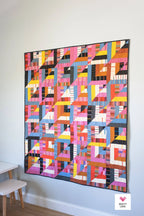 Colorful geometric quilt hanging on a white wall with a small stool in the corner. Lucky Log Cabin by Quilty Love