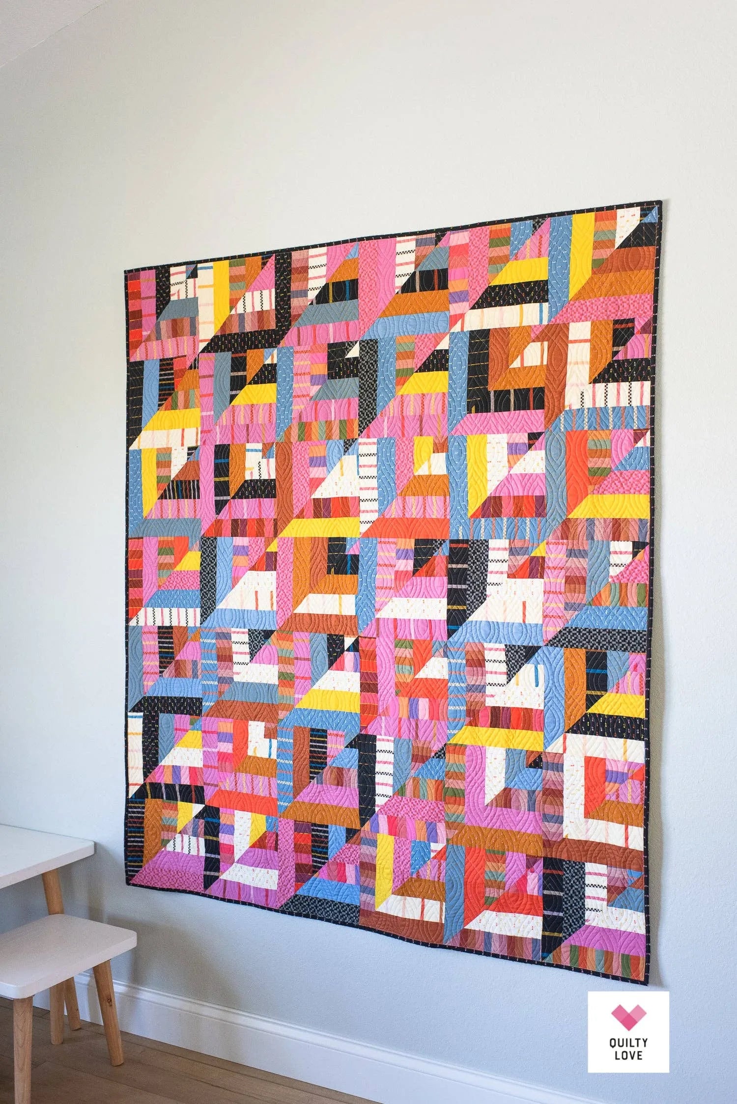 Colorful geometric quilt hanging on a white wall with a small stool in the corner. Lucky Log Cabin by Quilty Love