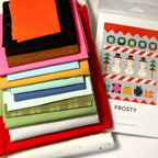 Fabric stack beside the pattern:
“Frosty quilt kit fabrics in bright holiday colors stacked beside the Modern Handcraft snowman quilt pattern.”