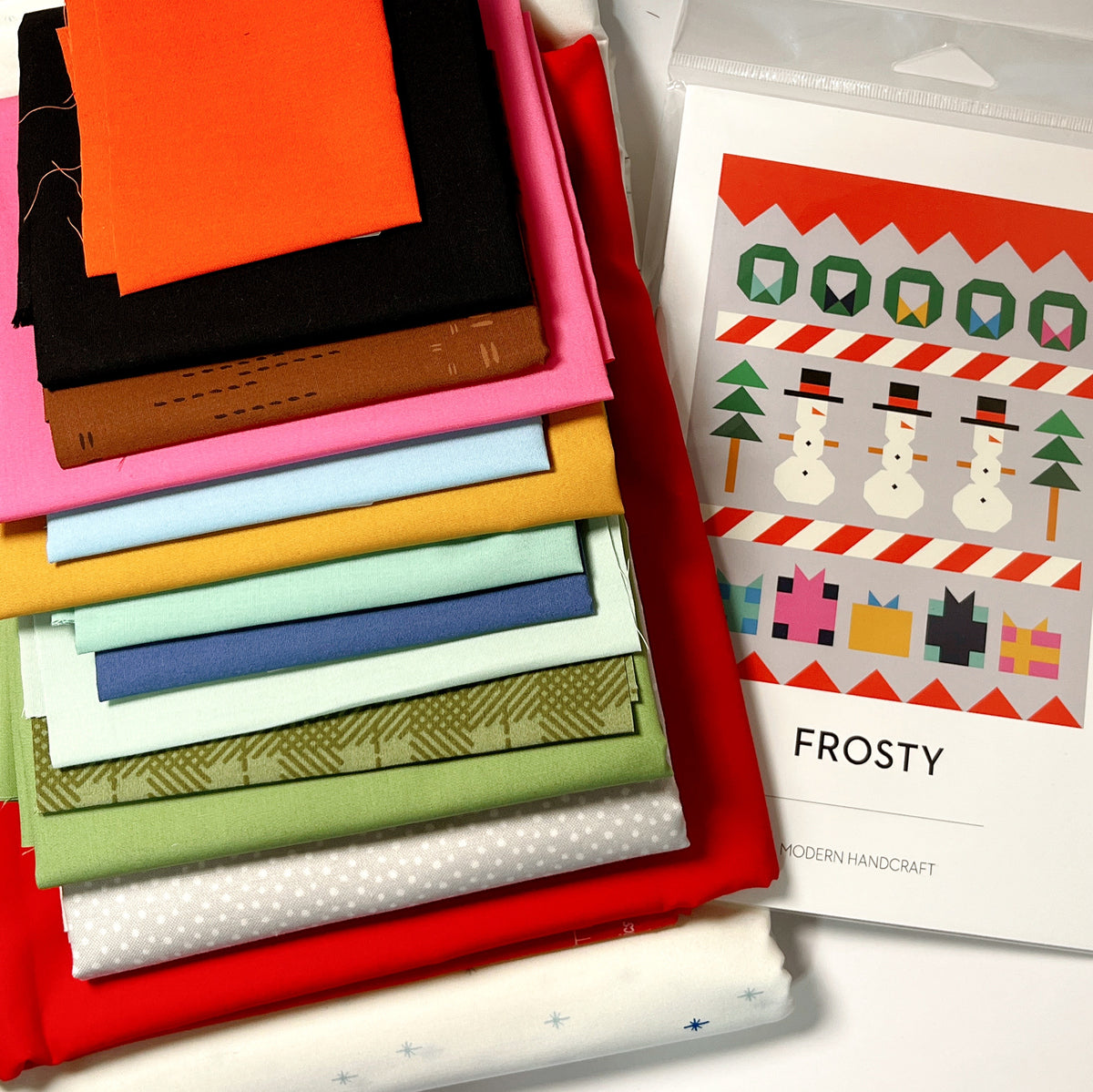 Fabric stack beside the pattern:
“Frosty quilt kit fabrics in bright holiday colors stacked beside the Modern Handcraft snowman quilt pattern.”