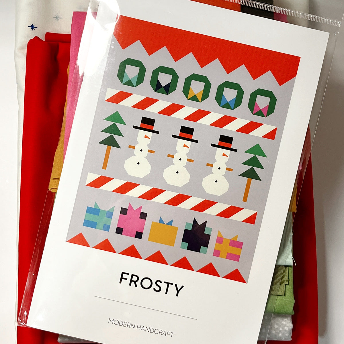 Pattern resting on the fabric bundle:
“Frosty quilt pattern resting on coordinating red, pink, green, and light-colored fabrics in the quilt kit.”