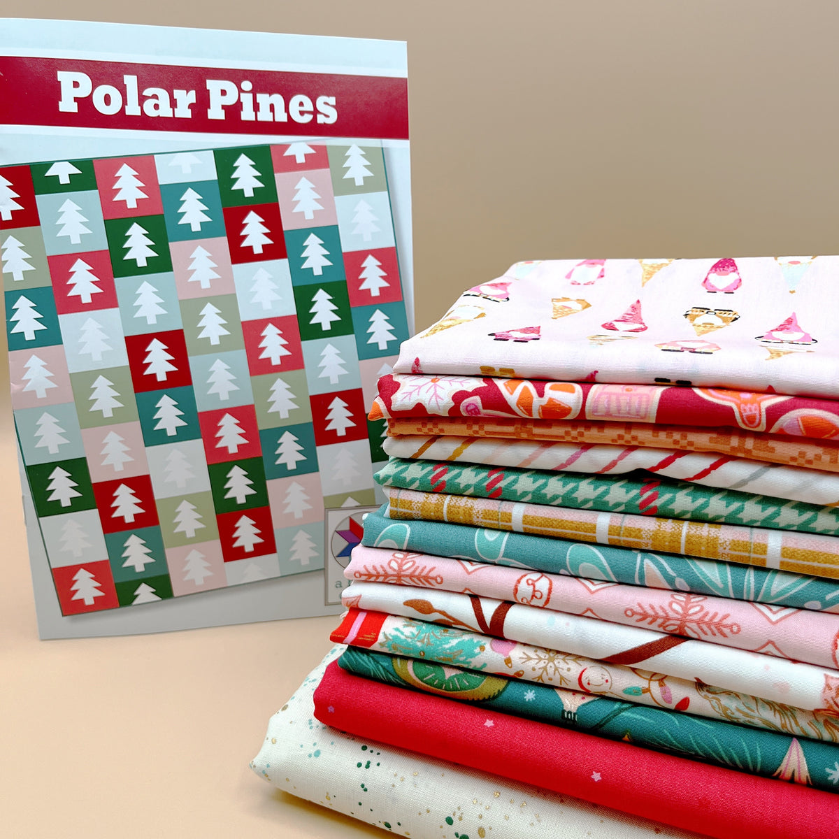 Polar Pines quilt pattern cover showing a grid of pine trees on red, green, teal, and pink blocks, next to a stack of folded Art Gallery Fabrics Christmas prints and a cream Ruby Star Speckled fabric with gold and green speckles.