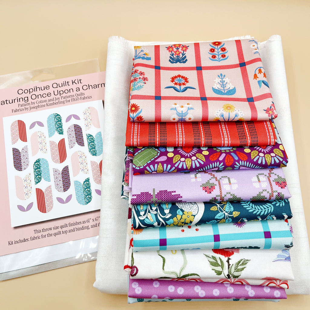 Fabric stack with kit label: “Once Upon a Charm printed fabrics stacked on white background fabric beside a Copihue quilt kit label showing the quilt design.”