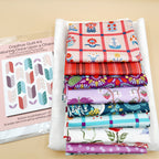 Fabric stack with kit label: “Once Upon a Charm printed fabrics stacked on white background fabric beside a Copihue quilt kit label showing the quilt design.”