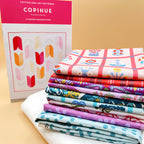 Pattern standing beside fabric stack: “Copihue pattern booklet beside stacked Once Upon a Charm fabrics in pink, red, purple, and teal on white background fabric.”