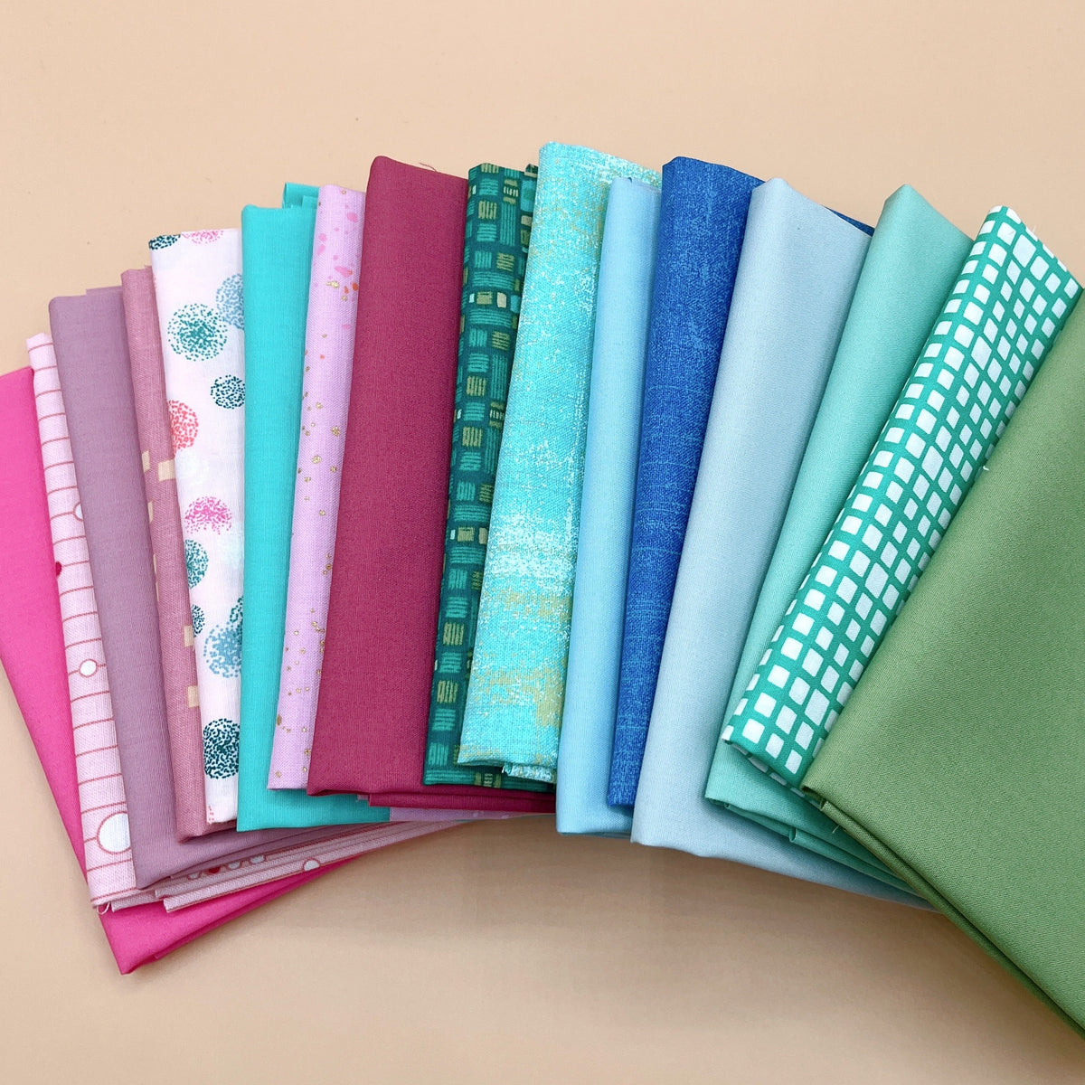 Sixteen folded fat quarters fanned out in a curve on a peach background, running from hot pink, mauve, and plum through pale pinks and speckled prints, then teal, aqua, and blue, ending in a teal and white grid print and olive green.