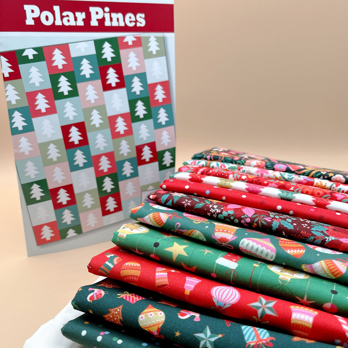 Polar Pines quilt pattern cover showing a grid of white pine trees on red, green, teal, and pink blocks, next to a stack of folded Deck the Halls Christmas fabrics with ornaments, stars, and holly in red and green.