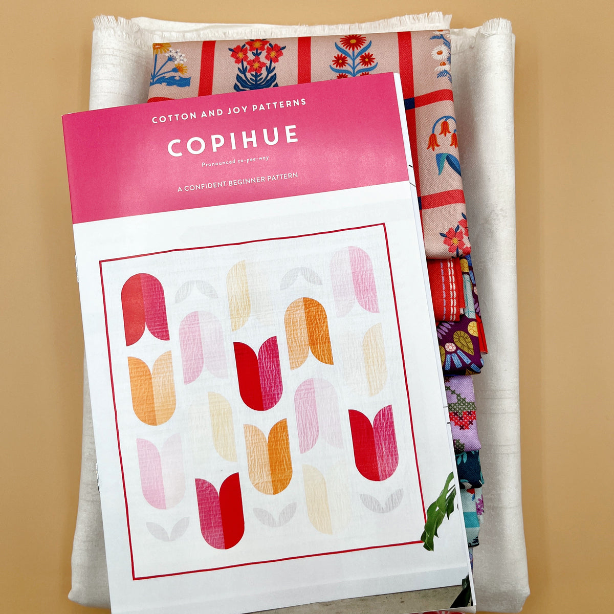 Pattern resting on fabric stack: “Cotton and Joy Copihue pattern booklet resting on folded Once Upon a Charm fabrics and white background fabric.”
