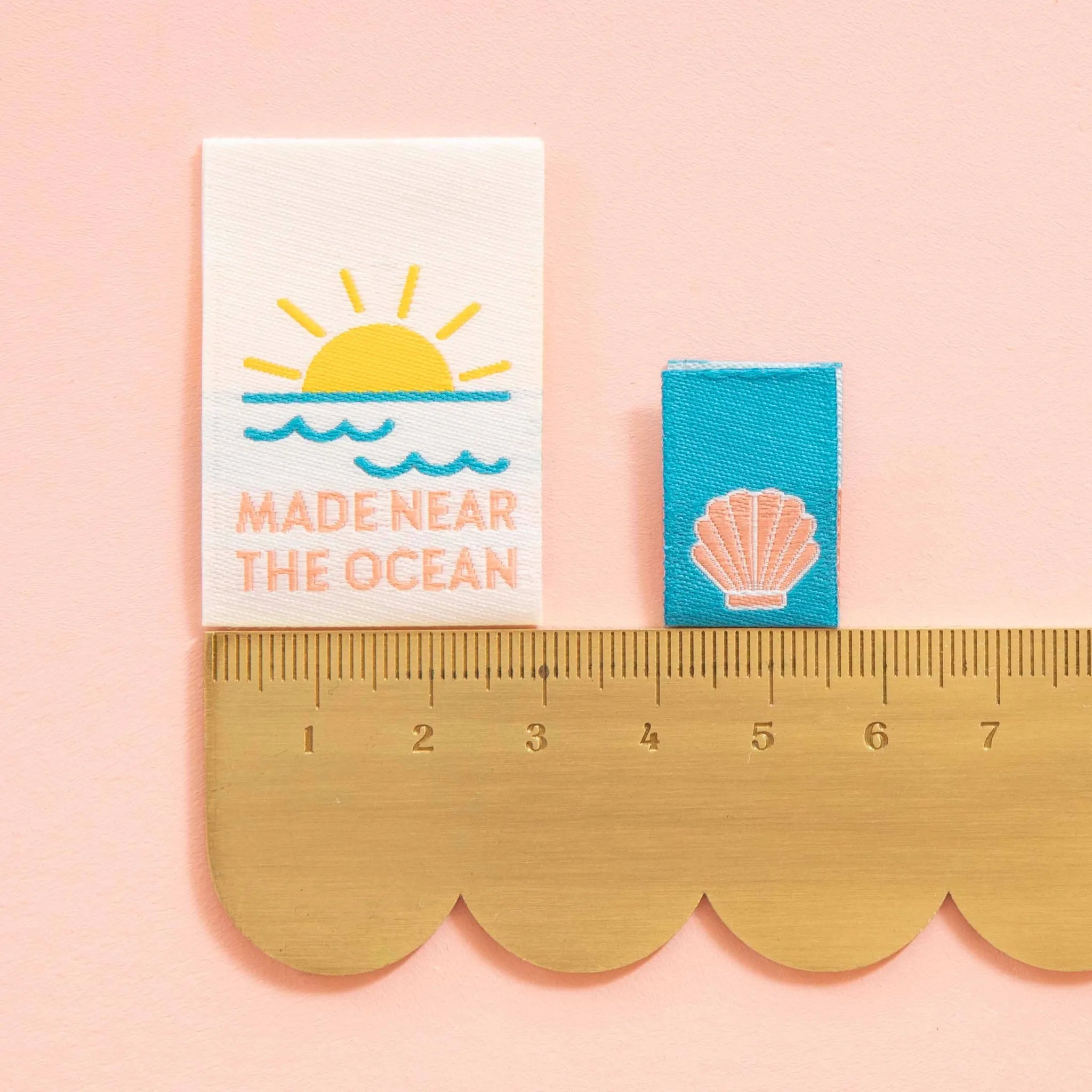 Sarah Hearts Woven Labels with ocean-themed design, blue ribbon, and gold ruler on pink background
